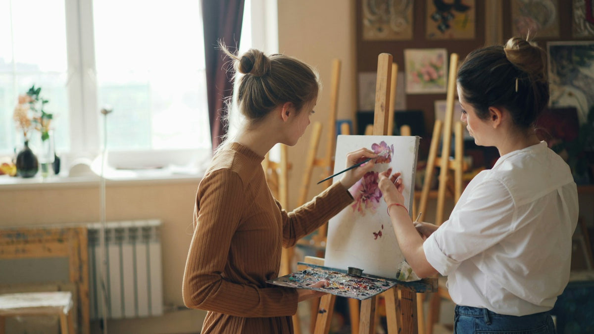 Two women painting together at easels in a bright art studio