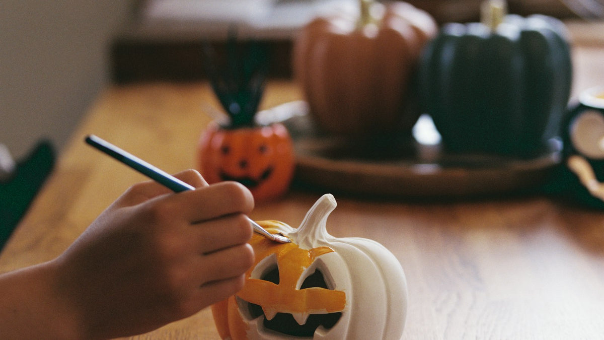 Hand painting a jack-o'-lantern face on a small white pumpkin at a wooden table with pumpkins behind