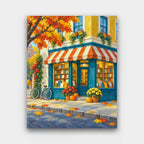 Bookshop on the Corner paint by numbers kit of a teal corner bookshop with a striped awning, pumpkins, mums and a bicycle in autumn