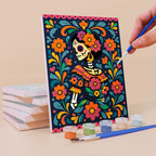 Painting a Día de Muertos mini canvas propped up beside numbered acrylic paints