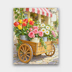 The Flower Cart paint by numbers kit of a wooden cart loaded with buckets of tulips, roses and daisies on a cobbled street