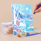 Painting a Winter Wonderland mini canvas propped up beside numbered acrylic paints